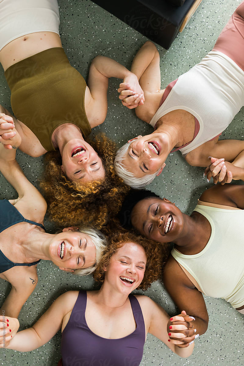 Above shot of five women from different background, holding hands and laughing in pilates studio